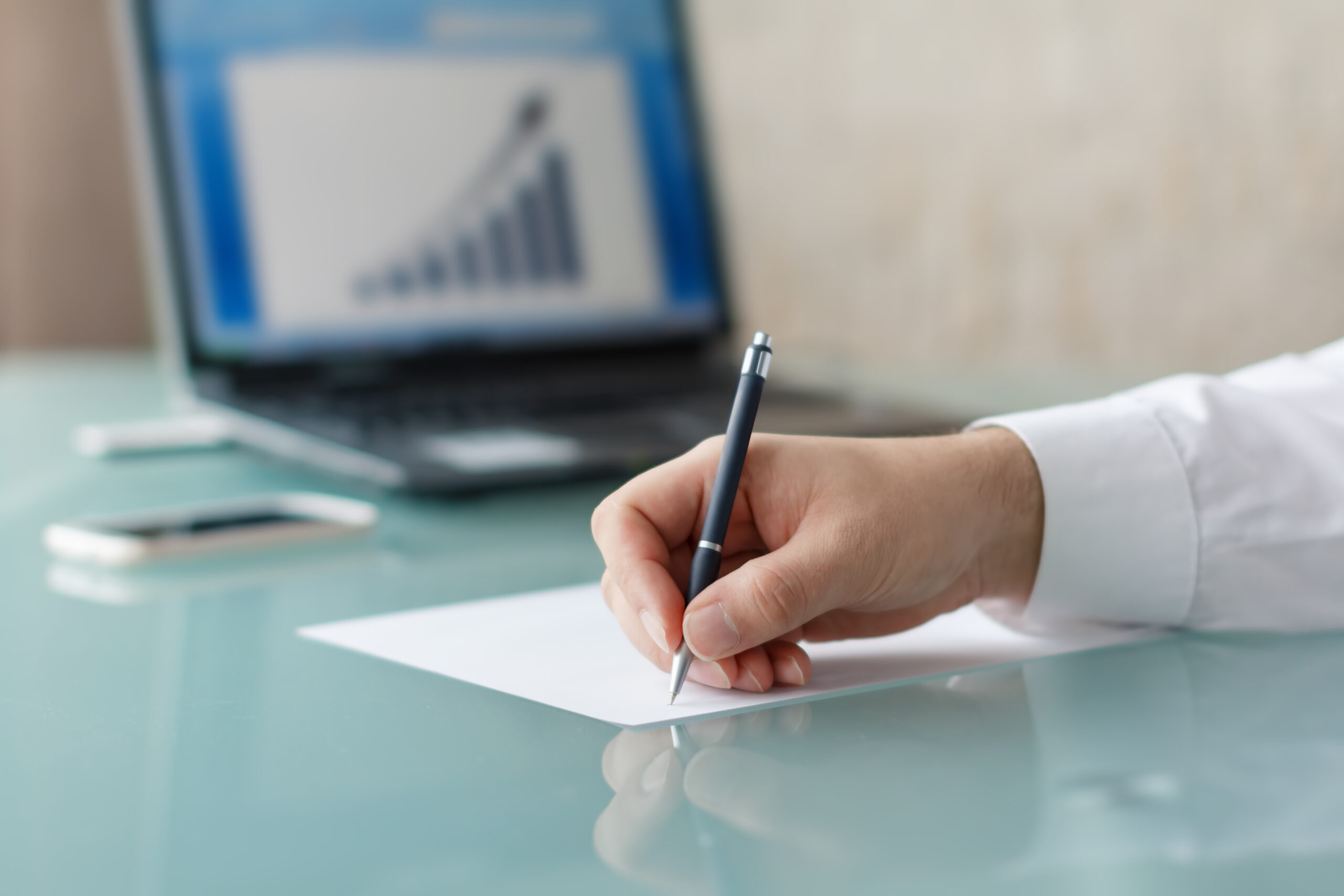 Hand holding a pen writing on paper with business analytics report on a laptop screen in the background.