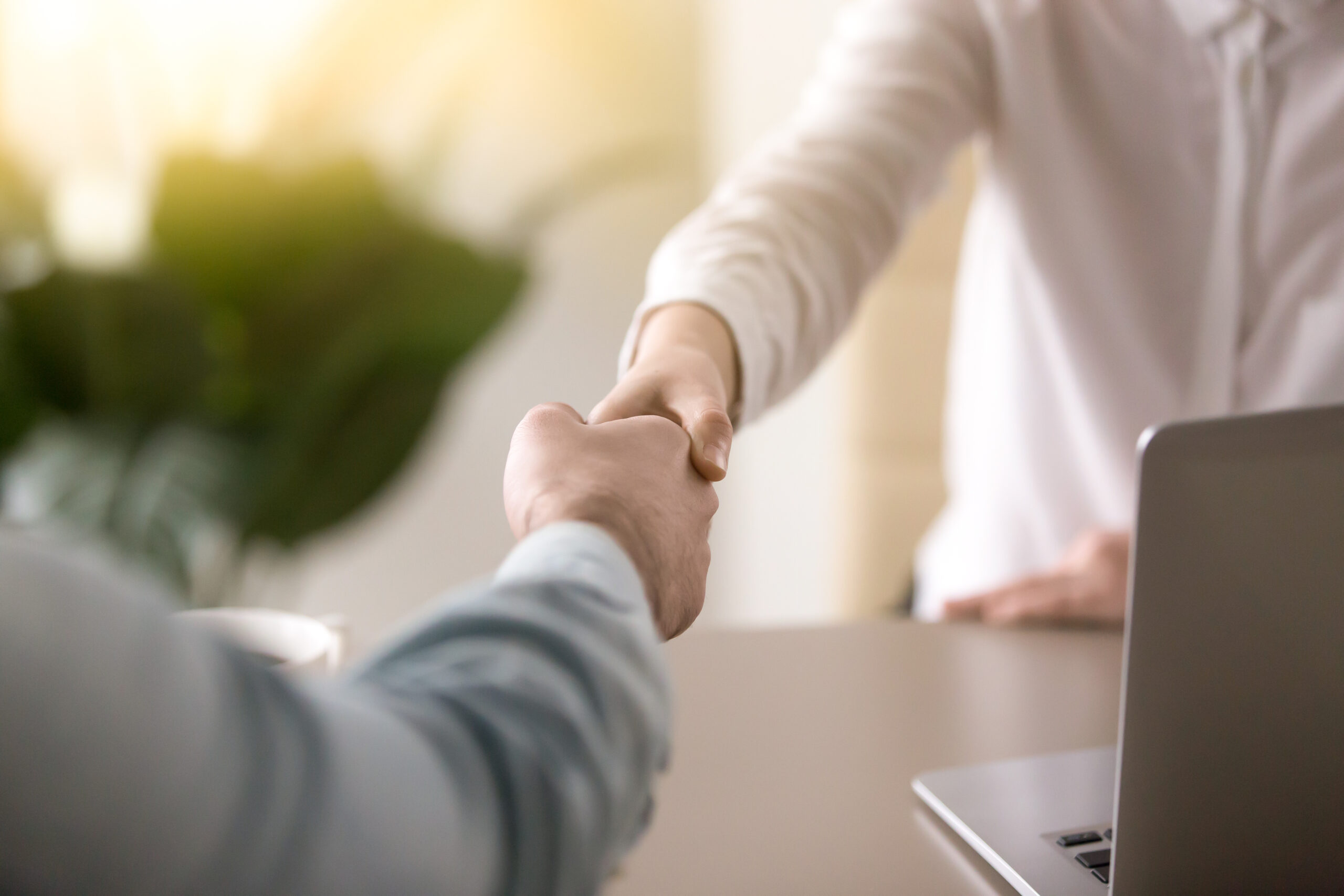 Two people shaking hands across a table with a laptop in the background.