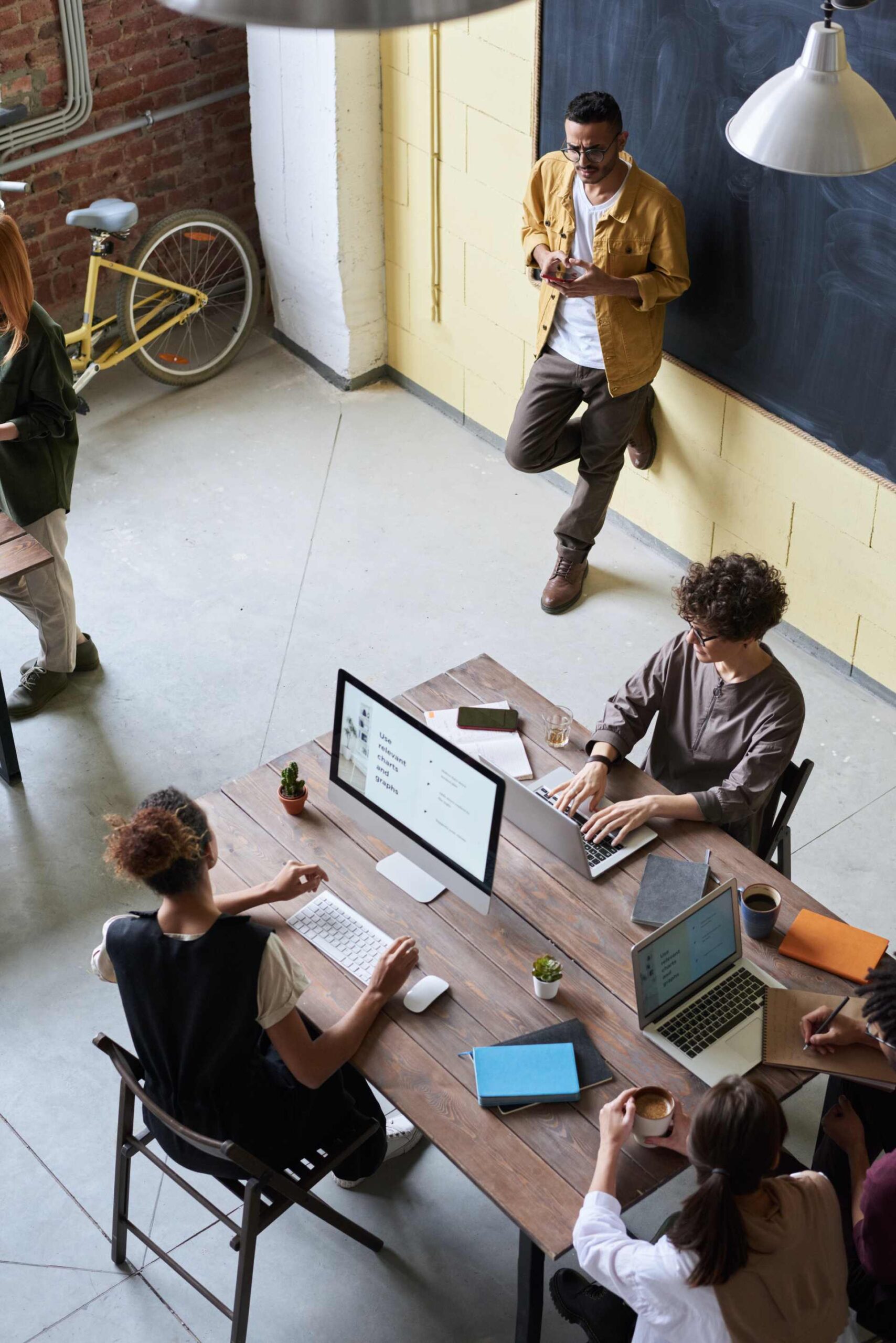 Team working collaboratively in a modern office, with individuals using laptops and desktops, while one person leans against the wall.