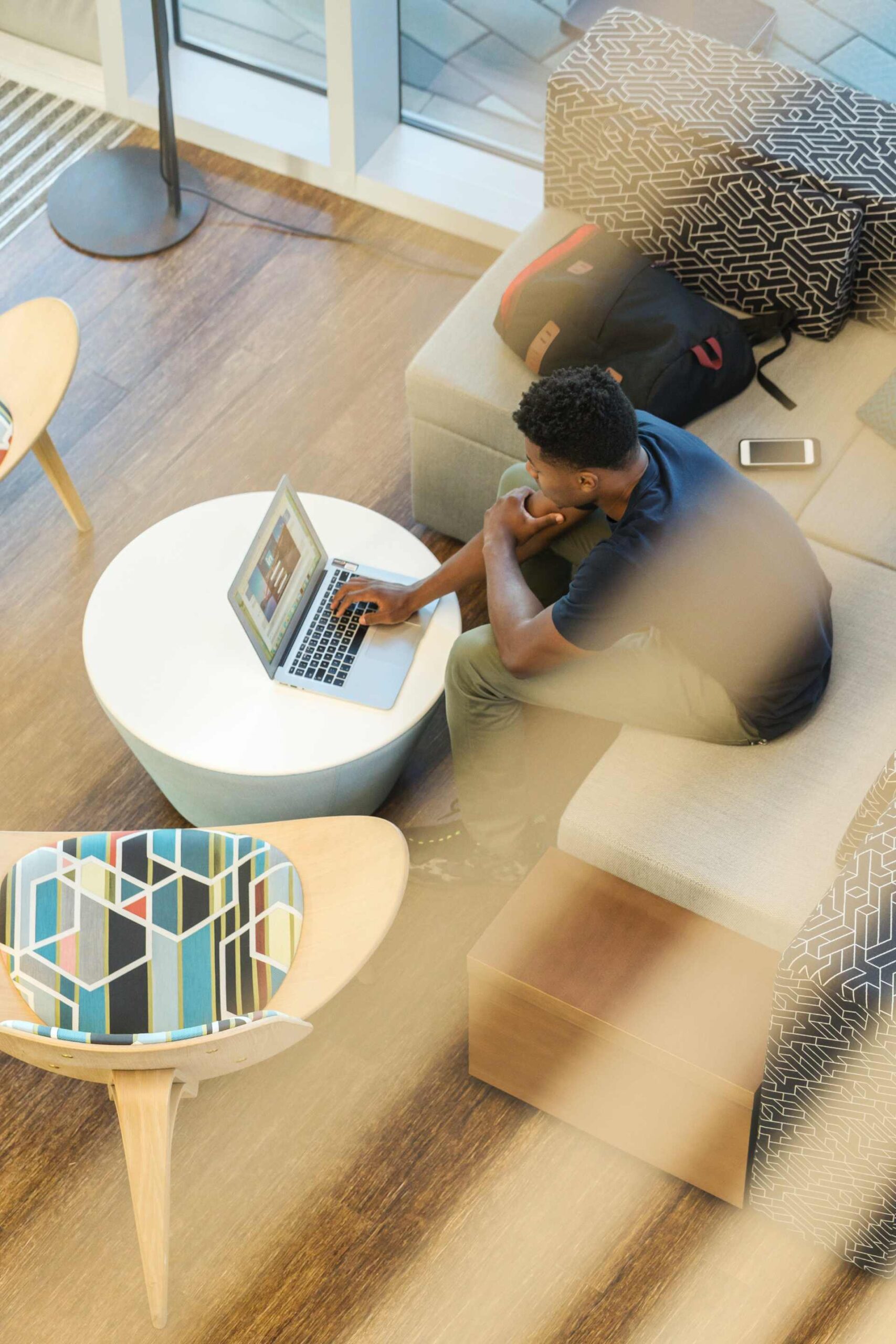 A student working on a laptop while sitting in a modern seating area with a backpack and phone nearby.